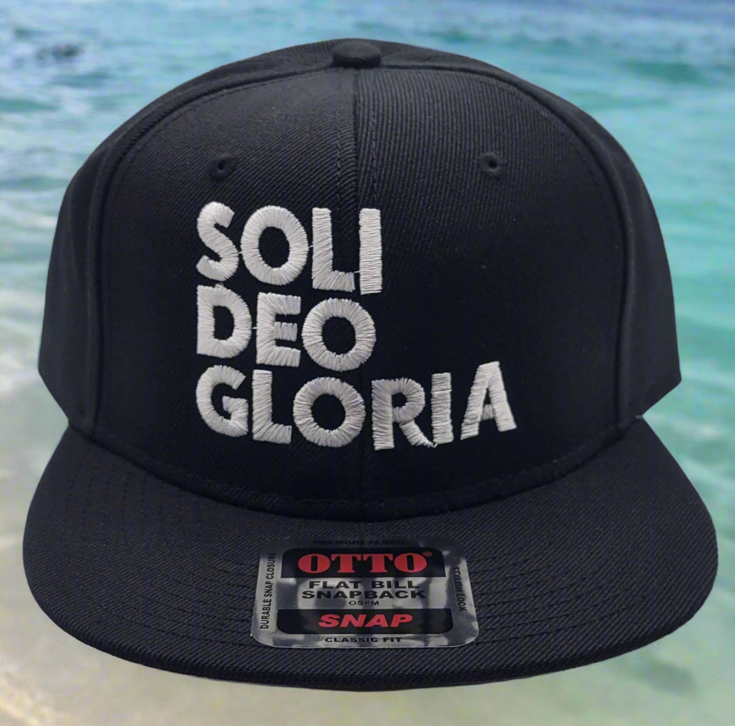 A black flat bill cap with the text 'SOLI DEO GLORIA' embroidered on the front, with a gray undervisor and the brand tag visible.