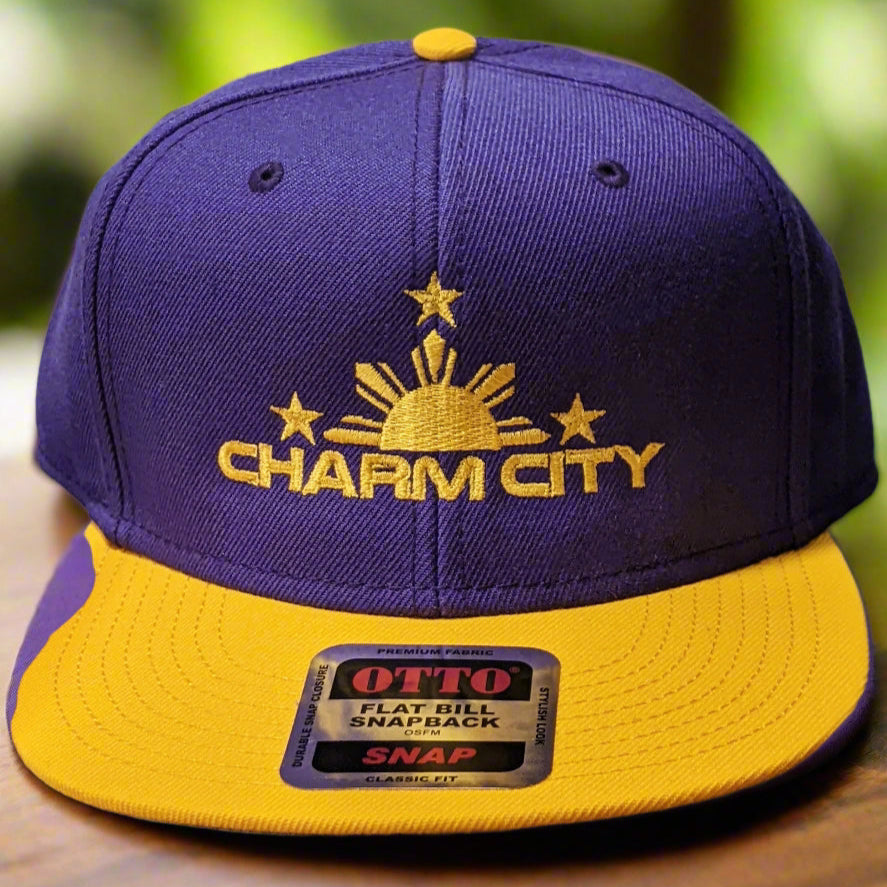 A purple and yellow flat bill snapback hat with 'Charm City' text and sun graphics embroidered on the front.